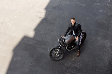 Obraz na płótnie Canvas Top view of a handsome rider man in black biker jacket and checkered shirt look to camera sit on classic style cafe racer motorcycle. Bike custom made in vintage garage. Brutal fun urban lifestyle.