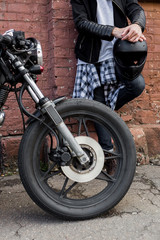 Closeup of a handsome rider biker guy hand with black helmet near brick wall and classic style cafe racer motorcycle. Bike custom made in vintage garage. Brutal fun urban lifestyle. Outdoor portrait.