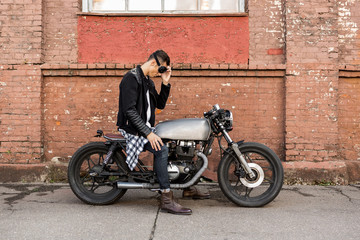 Fototapeta na wymiar Handsome rider biker man in black leather jacket and protection glasses sit on classic style cafe racer motorcycle. Bike custom made in vintage garage. Brutal fun urban lifestyle. Outdoor portrait.