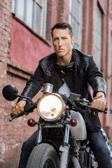 Fototapeta na wymiar Handsome rider biker man in black leather jacket, jeans, boots and gloves sit on classic style cafe racer motorcycle. Bike custom made in vintage garage. Brutal fun urban lifestyle. Outdoor portrait.