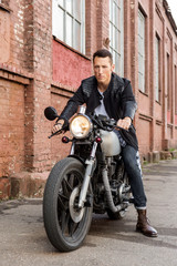 Fototapeta na wymiar Handsome rider biker guy in leather jacket sit on classic style cafe racer motorcycle and ready for long ride. Bike custom made in vintage garage. Brutal fun urban lifestyle. Outdoor portrait.