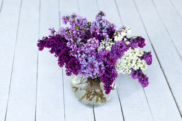 A bouquet of fresh lilac flowers in a glass vase on a wooden floor.