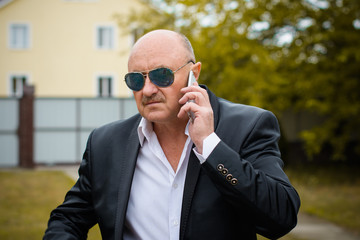 Portrait stylish mature men in white shirt and jacket and blue sunglasses using smartphone. Serving internet via his gadget. Old man using social network technology. Senior man talking on the phone