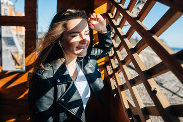 the magnificent girl with dark hair and a fine smile to look through a wooden window at the sea. She listens to music in white earphones and enjoys a sunny weather.