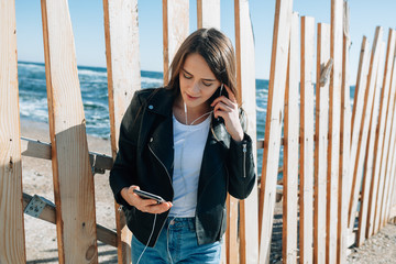 Lovely girl by the sea in white headphones. She has a black leather jacket and a white T-shirt. She is something on the phone.