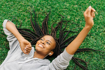 Above portrait of the joyful afro-american teenager laying on the grass while listening to music and moving hands.
