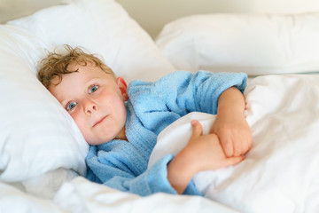 Little cute boy in blue pajamas with wet hair after bathing is lying on a huge snow-white pillow, wrapped in a blanket. He positively grimaces to the viewer.