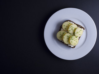 Danish open faced sandwich (smoerrebroed) with smoked cheese, sliced new potatoes and chives on dark rye bread. Placed on white plate on a black background.
