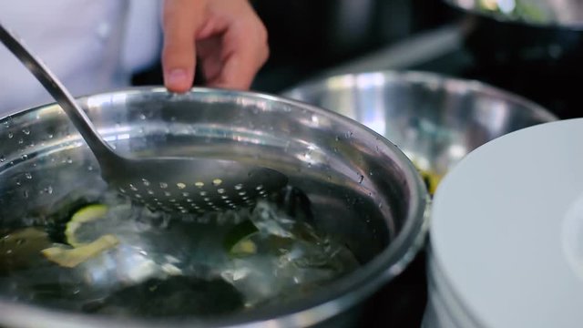 Cook catches skimmer sliced zucchini from the pan with water in a bowl.