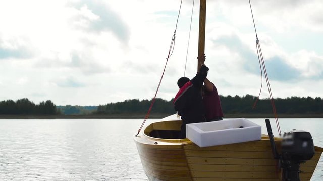 Mature Caucasian Master and his asian assistant working on wooden sailboat on river or lake. Smooth movement footage. They try to install mast. Middle shot.