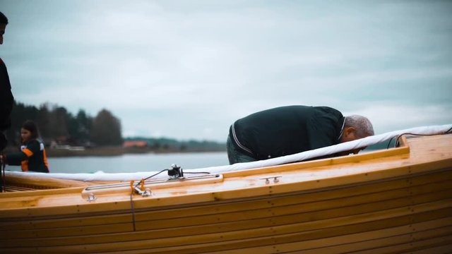 Mature Caucasian Master and his asian assistant working on wooden sailboat on river or lake. Smooth movement footage. They try to install mast. Close-up shot teal and orange.