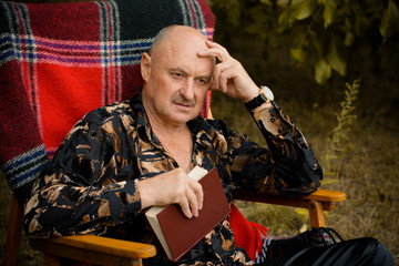 Portrait of old elegant man with gray hair reading a book in plaid armchair outside in park and thinking about his life