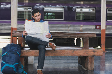 asian man with backpack sitting on platform at train station. backpacker or traveler look at map while waiting for train. journey, trip, travel concept