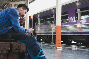 asian man with backpack sitting on platform at train station. backpacker or traveler with headphone look at mobile phone while waiting for train. journey, trip, travel concept