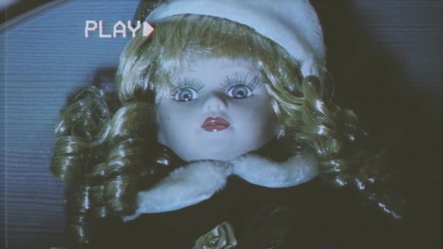 Fake VHS tape: a scary doll on top of a bed, in a bedroom, staring at the camera. Zoom in, we see her full creepy face.
