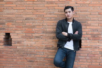 young asian man wearing black jacket and blue jeans standing against old orange brick wall
