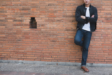 Fototapeta na wymiar young asian man wearing black jacket and blue jeans standing against old orange brick wall