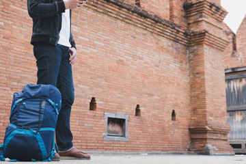 young traveler, asian man wearing black jacket and blue jeans standing near old orange brick wall with mobile smart phone and backpack