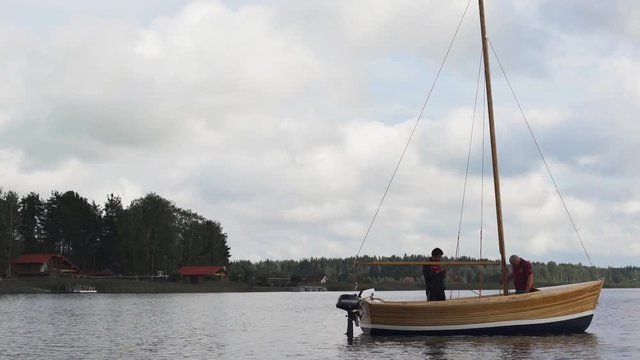 Mature Caucasian Master and his asian assistant working on wooden sailboat on river or lake. Smooth movement footage. They try to install mast. Wide shot