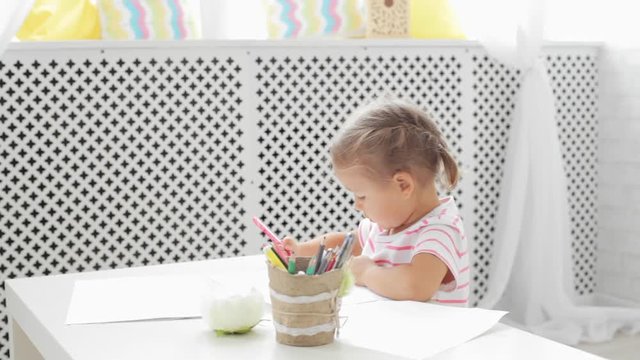 Cute preschool girl sitting by the white table focused on drawing something.