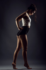 Fototapeta na wymiar Portrait of a beautiful slender girl athlete with updo hair dancing barefoot under studio searchlights wearing black high waist panties and white top. Isolated on dark background
