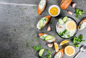 Traditional Caucasian and Greek food. Sauce tzatziki with ingredients - cucumber, lemon, parsley, dill, garlic. On a dark stone table. With sandwiches and baguette. Top view copy space