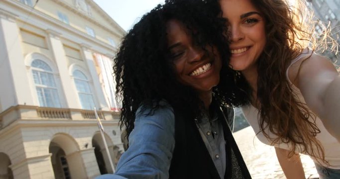 Selfie of two happy multicultural gilrfriends making duck face, kissing in the cheeks while walking the street.