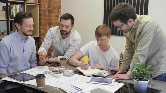 Red-haired man wrote in pencil on a sheet, and the other three look. In creative office, four people hold a meeting to address the issue of choosing the color of the interior. Funny guys with a beard