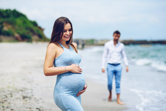 A beautiful pregnant woman in a blue tight dress touching her belly with love and care in the foreground and her courageous husband in the background on the seashore.