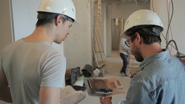 Two builders reviewing interior layout before starting finishing works indoors. They hold in their hands colored 3d images of future interior, pointing at its details with pen and talking actively