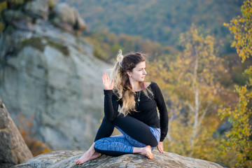 Calm young woman is practicing yoga and doing asana Arha Matsyendrasana on the top of the high rocky mountain in the evening. Autumn forests, rocks and hills on the background