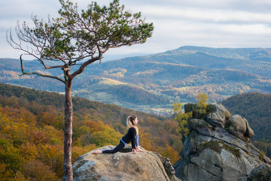 Active fit woman is practicing yoga on the top of the mountain near big tree. Autumn forests, rocks and hills on the background