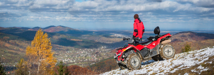 Panorama picture. Off-road vehicle and man near him on a snowy mountain top under the blue cloudy sky on a blurred background of autumn mountains and the town in the valley with copy space © anatoliy_gleb