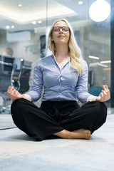 Young stressed hispanic business woman doing yoga inside office building, sitting in lotus position with hands on knees. Concept of long working hours and need of stress free break