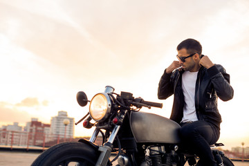 Handsome rider man with beard and mustache in black fashion sunglasses smoking cigaret and correct biker jacket sit on classic style cafe racer motorbike at sunset. Brutal fun urban lifestyle. - 164361789