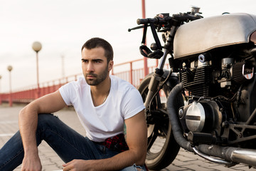 Fototapeta na wymiar Rider man with beard and mustache in clear white t-shirt sit near his classic style biker cafe racer motorcycle and look to the side. Bike custom made in vintage garage. Brutal fun urban lifestyle.