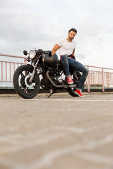 Handsome rider man with beard and mustache in red sneakers sit on classic style biker cafe racer motorcycle look to the camera. Bike custom made in vintage garage. Brutal fun urban lifestyle.