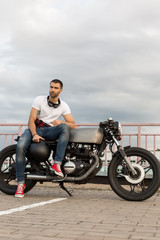 Fototapeta na wymiar Handsome rider man with a beard and mustache sit on classic style biker cafe racer motorcycle and and wait girlfriend. Bike custom made in vintage garage. Brutal fun urban lifestyle. Outdoor portrait.