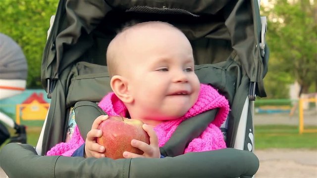 Baby girl sits in a baby stroller and eats an apple