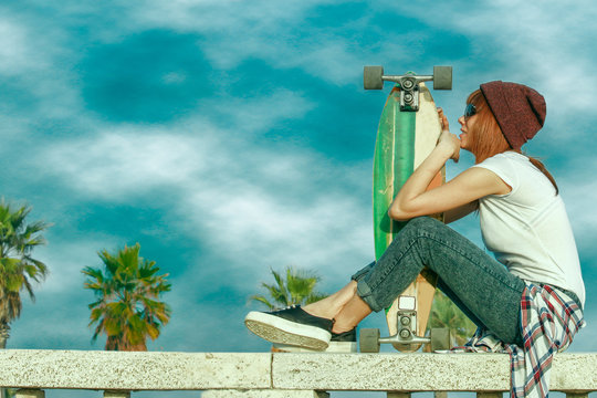 skater girl in jeans sitting and holding skateboard on a sunny day with cloudy sky. deep blue sky and saturated colors