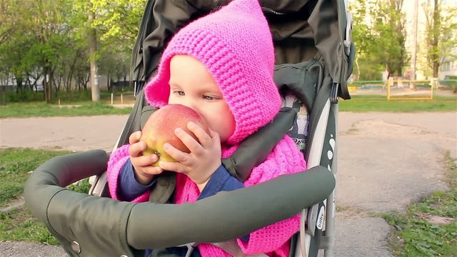 Baby girl sits in a baby stroller and eats an apple