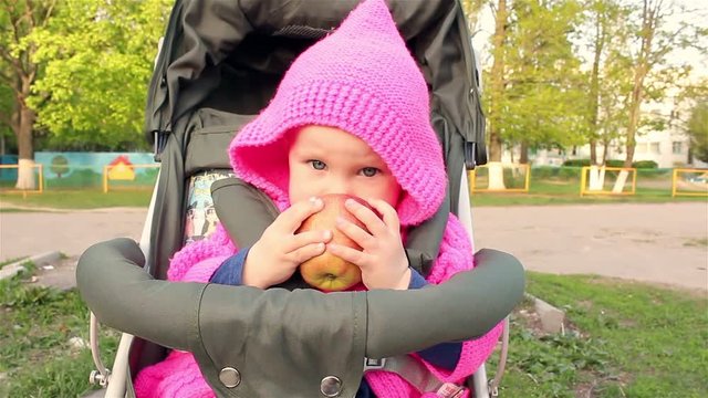 Baby girl sits in a baby stroller and eats an apple