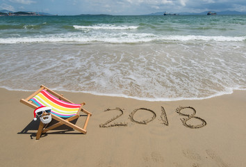 2018 written in sand write on tropical beach.Deck chair with Santa Claus sunglasses