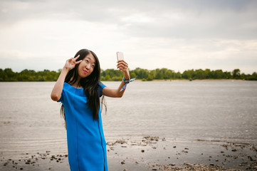 Young attractive Asian woman dressed in a blue dress, with long dark hair. Makes emotional selfie portrait photo on smartphone, on background of pond river outdoors by summer day