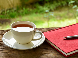 White cup of hot tea on wooden board with red notebook and pencil background. Afternoon teatime in the garden. Selective focus.
