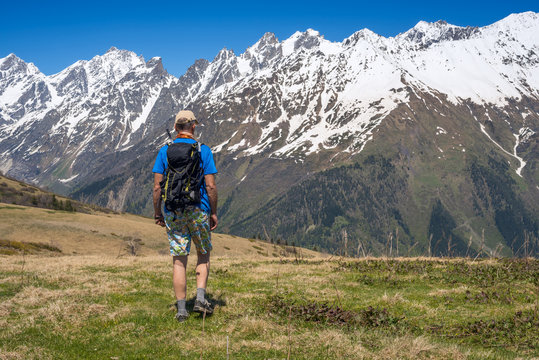 Man hiker goes along alpine meadow to the snow-capped mountain range