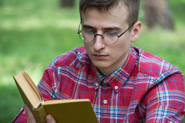 A student wearing glasses is reading a book in the park