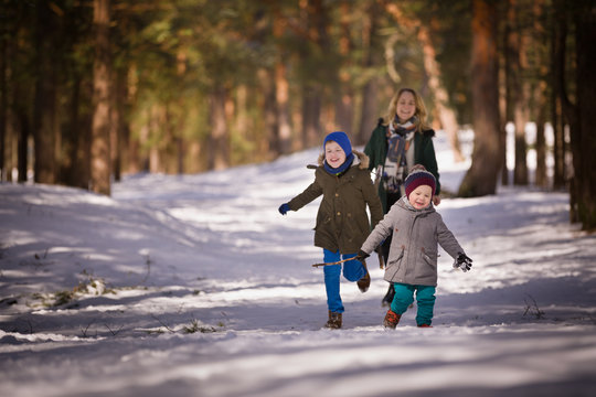 Happy family walking in a winter forest. Young woman, cute toddler boy and kid boy smiling and running on the snow on a sunny day.