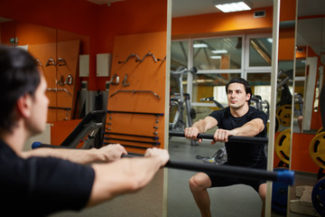 Young sporty man in the black sportwear with barbell doing squats in gym in front of the mirror.