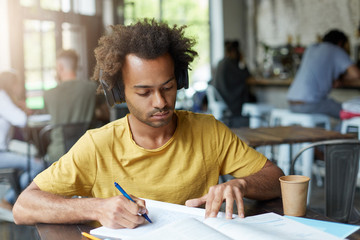 Fashionable African American university student doing homework on French at cafeteria, studying pronunciation and spelling, listening to audio tasks using headphones while learning new words